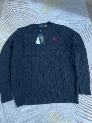 Svart kabelstickad tröja Polo Ralph Lauren - Kabelstickad svart tröja från Polo Ralph Lauren med rund halsringning och röd broderad logga på bröstet. Tröjan har ribbade muddar vid ärmslut och nederkant. Perfekt för dig som gillar klassisk och stilren design.
