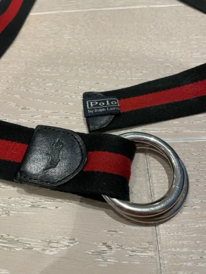 Randigt bälte från Polo Ralph Lauren - Snyggt bälte från Polo Ralph Lauren med svart och röd randig design. Bältet har dubbla silverfärgade D-ringar och detaljer i svart skinn med Polo-logga. Perfekt accessoar för att lyfta din outfit. Org pris: 800