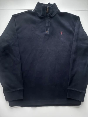 Marinblå half zip från Polo Ralph Lauren - Klassisk marinblå tröja från Polo Ralph Lauren med half zip och hög krage. Tröjan har lång ärm och den ikoniska röda loggan broderad på bröstet. Tillverkad i mjuk bomull för skön känsla och stilren look.
