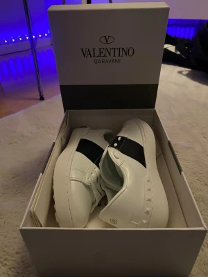 Valentino Garavani skor  - Snygga vita sneakers från Valentino Garavani med svart band över mitten och nitar på sidan. Skorna har klassisk låg profil, vita snören och är tillverkade i läder. Perfekta för dig som vill ha en clean och exklusiv look. Helt oanvända 