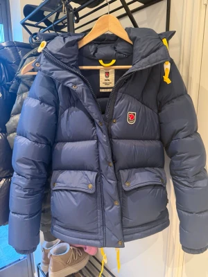 Blå dunjacka från Fjällräven XS  - Snygg blå dunjacka från Fjällräven med huva, gula detaljer och två stora fickor framtill. Jackan har en klassisk puffer-modell och är perfekt för kalla vinterdagar. Fjällräven-logga på bröstet och dragkedja med tryckknappar. Materialet är slitstarkt och isolerande. Inköpt i höstas så endast använden en säsong. Finns ett litet litet skrapmärke på ena fickan men som inte syns i det stora hela. 
