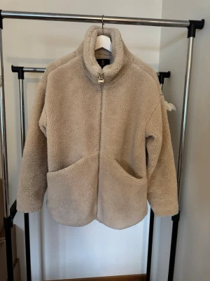 Teddyjacka Pernilla Wahlgren - Mysig beige teddyjacka från Pernilla Wahlgren med hög krage och dragkedja med PW-logga. Jackan har två stora fickor framtill och är gjord i fluffigt teddy-material som håller dig varm och stylish under kyliga dagar.