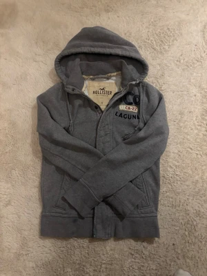 Hollister hoodie - Snygg grå hoodie från Hollister med dragkedja och broderade detaljer på bröstet där det står HCO, CA-22 och LAGUNA. Hoodien har huva med dragsko och två fickor framtill. Perfekt för en avslappnad och cool stil.