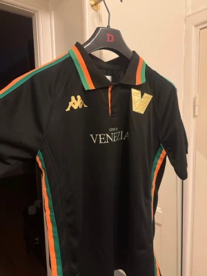 Venezia FC svart matchtröja Kappa - Snygg svart matchtröja från Venezia FC med guldiga Kappa-loggor och klubbmärke. Tröjan har krage med orange och gröna detaljer samt ränder längs sidorna. Tillverkad i lätt och ventilerande funktionsmaterial, perfekt för fotboll eller träning.