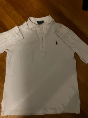 Vit pikétröja från Polo Ralph Lauren - Klassisk vit pikétröja från Polo Ralph Lauren med krage, knappar framtill och den ikoniska broderade loggan på bröstet. Tröjan har korta ärmar och är tillverkad i mjuk bomull, perfekt för en clean och stilren look.