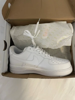 Nike Air Force 1 vita sneakers - Klassiska Nike Air Force 1 sneakers i helvitt läder med vit sula och vita skosnören. Ikonisk design med perforerad tå, låg profil och den välkända Swoosh-loggan på sidan. Perfekta för dig som vill ha en clean och tidlös look.