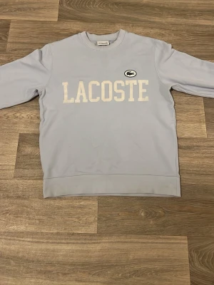 Ljusblå sweatshirt från Lacoste - Ljusblå stickad tröja från Lacoste med stort vitt tryck över bröstet och det klassiska krokodilmärket i svart och vitt. Tröjan har rund halsringning och långa ärmar. Perfekt för en avslappnad och stilren look.