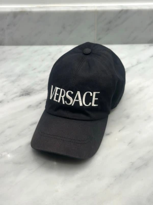 Svart keps från Versace - Svart keps från Versace med böjd skärm och justerbar rem baktill. Kepsen har en klassisk design med vit broderad logotyp framtill. Tillverkad i bomullstyg som ger en stilren och sportig look.