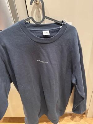 Mörkblå sweatshirt från No Nationality - Säljer en stilren mörkblå sweatshirt från No Nationality med vit text på bröstet. Tröjan har rund halsringning och långa ärmar. Tillverkad i mjuk bomull för en bekväm känsla och avslappnad passform.