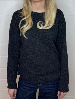 Mörkgrå stickad tröja - Mysig mörkgrå stickad tröja med rund halsringning och långa ärmar. Tröjan har en enkel, klassisk design och är tillverkad i ett mjukt material som passar perfekt till kalla dagar. Ribbstickade muddar vid ärmslut och nederkant.