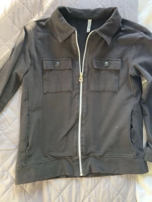 Svart overshirt med dragkedja - Stilren svart overshirt med vit dragkedja framtill, två bröstfickor med knappar och klassisk krage. Långärmad modell med knappdetaljer vid ärmsluten. Perfekt för lager på lager och enkel att matcha till olika outfits.