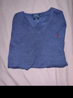 Blå stickad tröja från Polo Ralph Lauren - Snygg blå stickad tröja från Polo Ralph Lauren med V-ringning och klassisk röd logga broderad på bröstet. Tröjan har en enkel och stilren design, perfekt att matcha med jeans eller chinos. Materialet känns mjukt och bekvämt.