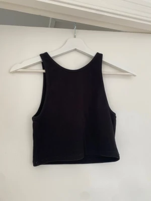 Svart ribbad croppad topp - Säljer en svart croppad linnetopp i ribbat material. Toppen har breda axelband och är ärmlös med en tight passform. Perfekt att styla med höga jeans eller kjol för en clean och enkel look.