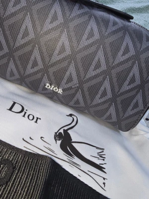 Grå axelväska från CD - Snygg grå axelväska från Dior med geometriskt mönster i olika nyanser av grått och silverfärgad logga framtill. Väskan har en bred axelrem med 'Christian Dior' broderat. Använd få gånger. Köpt för 1500 kr, säljer den för 900 kr
