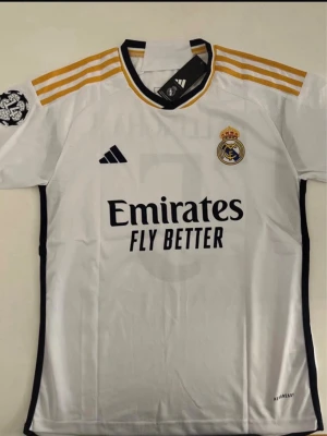 Sprillans Ny Real Madrid Matchtröja Bellingham #5 - Snygg vit Real Madrid matchtröja från Adidas med gula ränder på axlarna och klubbmärke på bröstet. Tryck med Bellingham och nummer 5 på ryggen. Tillverkad i lätt och ventilerande material, perfekt för fotboll eller träning.