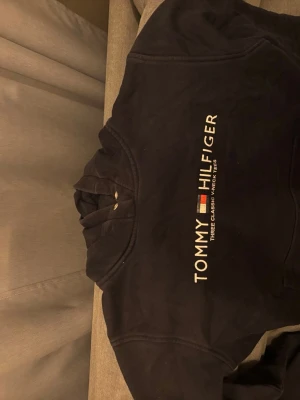 Svart hoodie från Tommy Hilfiger - Snygg svart hoodie från Tommy Hilfiger med klassisk logga och text på bröstet i vitt, rött och blått. Hoodien har huva och är tillverkad i mjukt bomullsmaterial. Perfekt för en avslappnad och trendig stil.