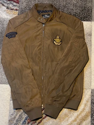 Brun bomberjacka från Morris - Snygg brun bomberjacka från Morris med patch på ärmen och broderad logga på bröstet. Jackan har ribbade muddar, ståkrage med knapp och hel dragkedja framtill. Fodret är mönstrat i blått och vitt för extra stilpoäng. Perfekt för dig som vill ha en klassisk men trendig look. Finns lite defekter med inget som man lägger märke till vid användning.
