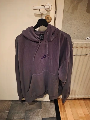 Lila hoodie från Adidas - Säljer en lila hoodie från Adidas med klassisk huva och dragsnören. Hoodien har en stor magficka framtill och Adidas-logga broderad på bröstet. Tillverkad i mjukt bomullsmaterial som känns skönt mot huden. Perfekt för en avslappnad och sportig stil.