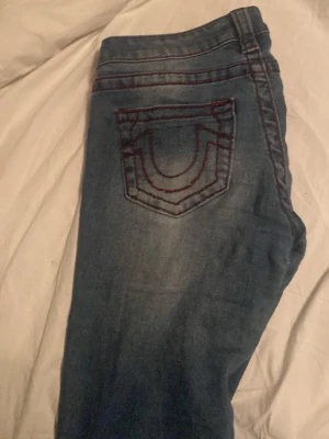 Blå True Religion jeans med röd söm - Säljer ett par blå jeans från True Religion med ikonisk röd kontrastsöm och klassisk fickdesign bak. Jeansen har en snygg tvättad look och silverfärgade knappar. Perfekta för dig som vill ha en streetig och trendig stil.