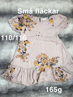 Ljusrosa blommig klänning Pompdelux - Söt ljusrosa klänning från pompdelux med volangkant nedtill och korta puffärmar. Klänningen har ett blommigt mönster. Perfekt för varma dagar och har en avslappnad passform.🌸 Små fläckar. Storlek 110/116