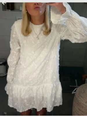 Vit blommig kortklänning med volang - Superfin vit kortklänning med blommigt mönster och volang nertill. Klänningen har långa ärmar och är tillverkad i ett lätt, luftigt material. Perfekt för sommarens alla tillfällen.