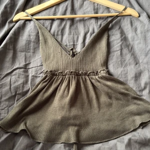 Grön stickad linnetopp med snörning - Supersöt mörkgrön stickad linnetopp med smala axelband och v-ringning. Toppen har ribbad överdel, volangkant i midjan och snörning i ryggen för en snygg passform. Perfekt för varma dagar och enkel att styla med jeans eller kjol.