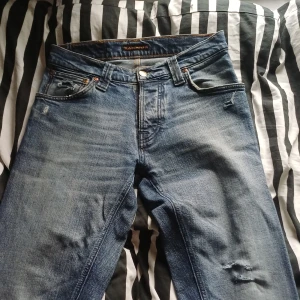 Blå Nudie Jeans, W30 L34, slitna detaljer - Säljer ett par blå jeans från Nudie Jeans med snygga slitningar och distressed detaljer. Klassisk femficksmodell med orangea sömmar och knappgylf. Jeansen har en rak passform och är tillverkade i robust denim. Perfekta för en avslappnad och cool stil.