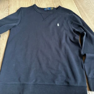 Svart tröja från Polo Ralph Lauren - Klassisk svart långärmad tröja från Polo Ralph Lauren med rund halsringning och broderad logga på bröstet. Tillverkad i mjuk bomull för en skön känsla och enkel stil. Perfekt för dig som gillar stilrena och tidlösa plagg.