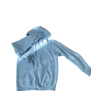 Grå rhinestone hoodie från BVALDI  - Grå zip-hoodie från BVALDI med huva och dragsko. Framsidan har ett stort motiv av en bevingad figur i RHINESTONE och glittrande detaljer, samt text i RHINESTONE på bröstet. Mjuk insida och två fickor framtill. Perfekt för dig som gillar statement-plagg/Laidback plagg