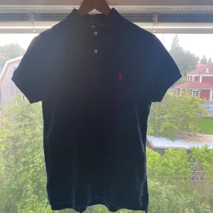 Mörkblå Polo Ralph Lauren piké S - Säljer en mörkblå pikétröja från Polo Ralph Lauren i storlek S. Klassisk modell med korta ärmar, två knappar vid kragen och röd broderad logga på bröstet. Tillverkad i 100% bomull och custom fit. Perfekt för en clean och stilren look.