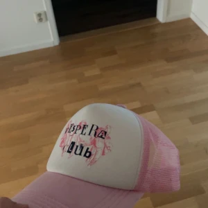 Rosa och vit truckerkeps vespera Club - Säljer en rosa och vit truckerkeps från vespera Club. Kepsen har vit frontpanel med tryck och rosa mesh på sidor och bak, samt justerbar  kepsen har en klassisk böjd skärm.