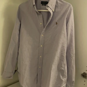 Ljuslila skjorta från Ralph Lauren - Säljer en klassisk ljuslila skjorta från Ralph Lauren med custom fit. Skjortan har button-down krage, broderad logga på bröstet och vita knappar. Tillverkad i bomull och har lång ärm. Perfekt för en clean och stilren look.