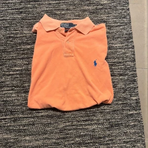 Orange pikétröja från Ralph Lauren - Snygg orange pikétröja från Polo Ralph Lauren i klassisk modell med krage och knappar. Tröjan har blå broderad logga på bröstet och är tillverkad i mjuk bomull. Perfekt för dig som vill ha en stilren och fräsch look.
