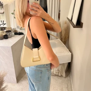Beige axelväska i skinnlook - Snygg beige axelväska i stilren, trekantig form med justerbart axelband och guldfärgad detalj framtill. Väskan har dragkedja upptill och är tillverkad i ett slätt material som liknar skinn. Perfekt storlek för det viktigaste och ger en clean vibe till din outfit.