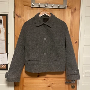 Grå ulljacka från 157, storlek M - Stilren grå jacka från 157 i ullmaterial med klassisk krage, stora framfickor och knäppning med tre knappar framtill. Jackan har raka, långa ärmar med knappdetalj vid ärmslut och en boxig, något croppad passform. Perfekt för kyliga dagar.