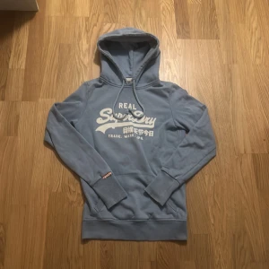 Blå hoodie från Superdry XS - Säljer en blå hoodie från Superdry i storlek XS. Tröjan har vit logga framtill, snörning i huvan och en klassisk känguruficka. Mjuk insida och ribbade muddar vid ärmslut och nederkant. Perfekt för en chill och avslappnad stil.