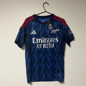 Arsenal bortatröja Adidas S blå - Arsenal bortatröja från Adidas i marinblått med blixtmönster och röda axelränder. Slim fit-modell. Tryckta loggor och sponsor på bröstet, klubbmärke på vänster sida. Perfekt för dig som vill sticka ut på läktaren eller planen.