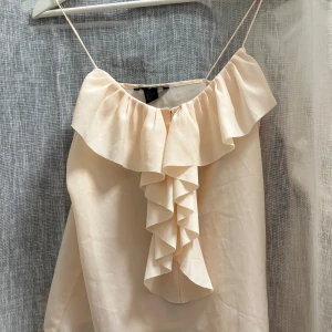Beige volangtop från H&M - Säljer en ljusbeige/rosa linnetopp från H&M med tunna axelband och volangdetaljer framtill. Toppen har en snygg omlottvolang längs halsringningen och ned över bröstet. Perfekt för sommarens varma dagar och enkel att styla till olika looks.