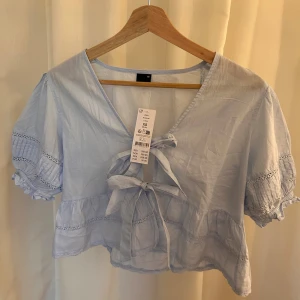 Ljusblå blus med knyt från Gina Tricot - Supersöt ljusblå blus från Gina Tricot med v-ringning och två knyt framtill. Den är helt ny med prislapp kvar! Blusen har korta puffärmar med spetsdetaljer och volang längs nederkanten. Perfekt för en fräsch och somrig look. Tillverkad i mjuk bomull. Nypris 399 kr.
