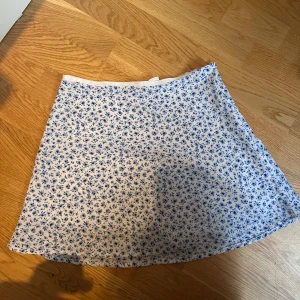 Blommig kortkjol  - Supersöt kortkjol från Divided i vitt med blått blommönster. Kjolen har en lätt A-linjeform och är gjord i ett mjukt bomullstyg som känns skönt mot huden. Perfekt för dig som gillar en fräsch och somrig stil med romantiska vibbar.