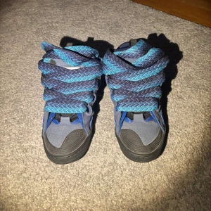 Lanvin curbs  - Unika sneakers i blått och svart med breda, mönstrade snören i blå och turkos. Skorna har svarta sulor, rund tå och detaljer i både matt och glansig blå finish. Ovandelen är i syntet och mockaliknande material, med coola vågformade detaljer på sidorna.