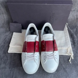 Valentino Garavani vita sneakers röd detalj - Snygga vita sneakers från Valentino Garavani med bred röd läderdetalj över mitten. Klassisk rund tå, vita snören och diskret logga på plösen. Skorna har en platt sula och är tillverkade i skinn för en lyxig känsla.