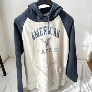 American Eagle hoodie i blått och vitt - Snygg hoodie från American Eagle i storlek L. Tröjan är vit framtill och baktill med marinblå ärmar och huva. Stor tryckt logga och örn på bröstet, samt känguruficka och snörning i huvan. Perfekt för en avslappnad och sportig look.