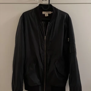 Svart bomberjacka från H&M L.O.G.G. - Svart bomberjacka från H&M L.O.G.G. med klassisk ribbad krage, dragkedja framtill och fickor med lock. Jackan har en extra ficka med dragkedja på ärmen och är tillverkad i ett slätt syntetmaterial. Perfekt för en stilren och urban look.