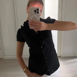Svart blus med volang från Zara Basic - Supersnygg svart blus från Zara Basic med korta puffärmar, volangdetaljer vid halsringningen och knappar framtill. Sitter figurnära och har en feminin v-ringning med knytband. Perfekt för dig som gillar stilrena men ändå unika plagg.