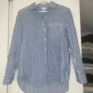 Blåvit randig skjorta i bomull - Snygg blåvit skjorta från H&M med vertikala ränder och klassisk krage. Skjortan har lång ärm, bröstficka och knappar framtill. Den har en fläck på ärmen men borde gå bort i tvätten har inte testat 