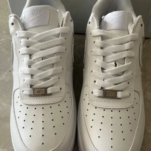 Nike Air Force 1 helvita sneakers - Klassiska Nike Air Force 1 sneakers i helvitt skinn med perforerad tå, rund tå och platt sula. Skorna har snörning och diskret Nike-logga på häl och plös. Perfekt för dig som vill ha en clean och tidlös look.