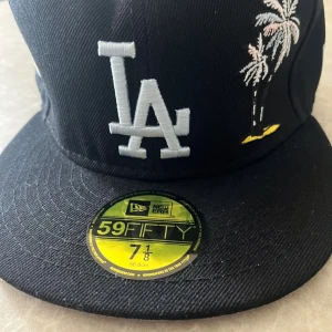 Svart LA Dodgers keps från New Era - Snygg svart 59FIFTY keps från New Era med LA Dodgers logga i vitt framtill, broderade palmer och ett gult inslag på sidan. På ena sidan finns ett Dodger Stadium 50th Anniversary märke och på baksidan ett vitt hjärta. Perfekt för dig som gillar streetstyle och baseball.