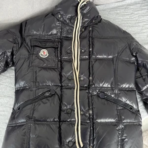 Svart Moncler dunjacka med dragkedjor - Säljer en svart, glansig dunjacka från Moncler med quiltad design och coola detaljer som snörning vid kragen och dragkedjor på fickorna. Jackan har en bröstficka med Moncler-logga, tryckknappar och vit insida med röd och marinblå rand. Perfekt för kalla dagar!