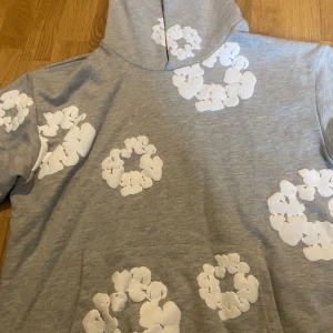 Grå hoodie med vita blommor Denim Tears - Grå hoodie från Denim Tears med stora vita blommor tryckta över hela tröjan. Tröjan har huva och en klassisk magficka framtill. Perfekt för dig som gillar streetwear med unik design och vill sticka ut lite extra.